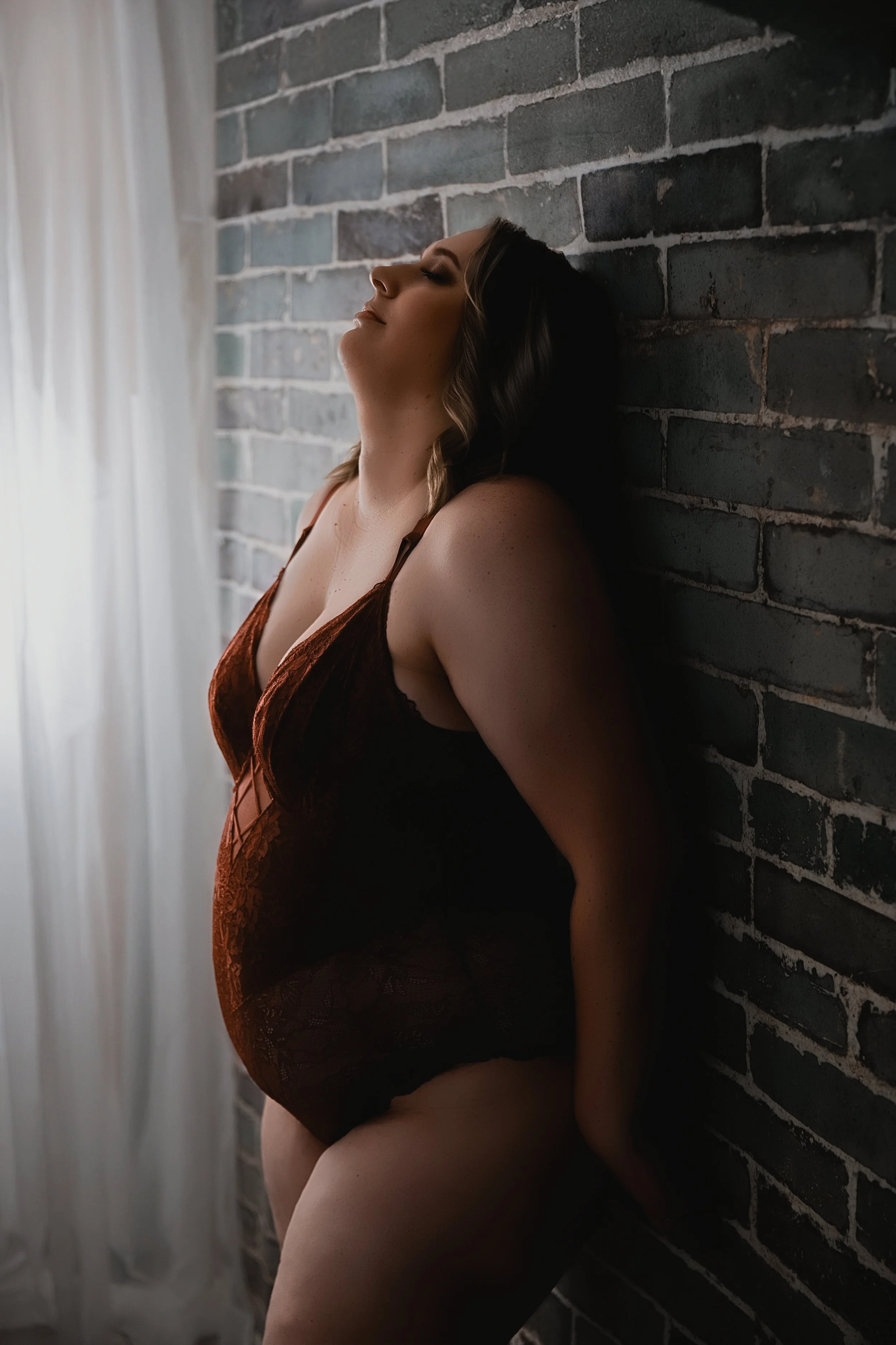 A woman wearing brown and black lace lingerie with her eyes closed, leaning against a brick wall with her head tilted back.