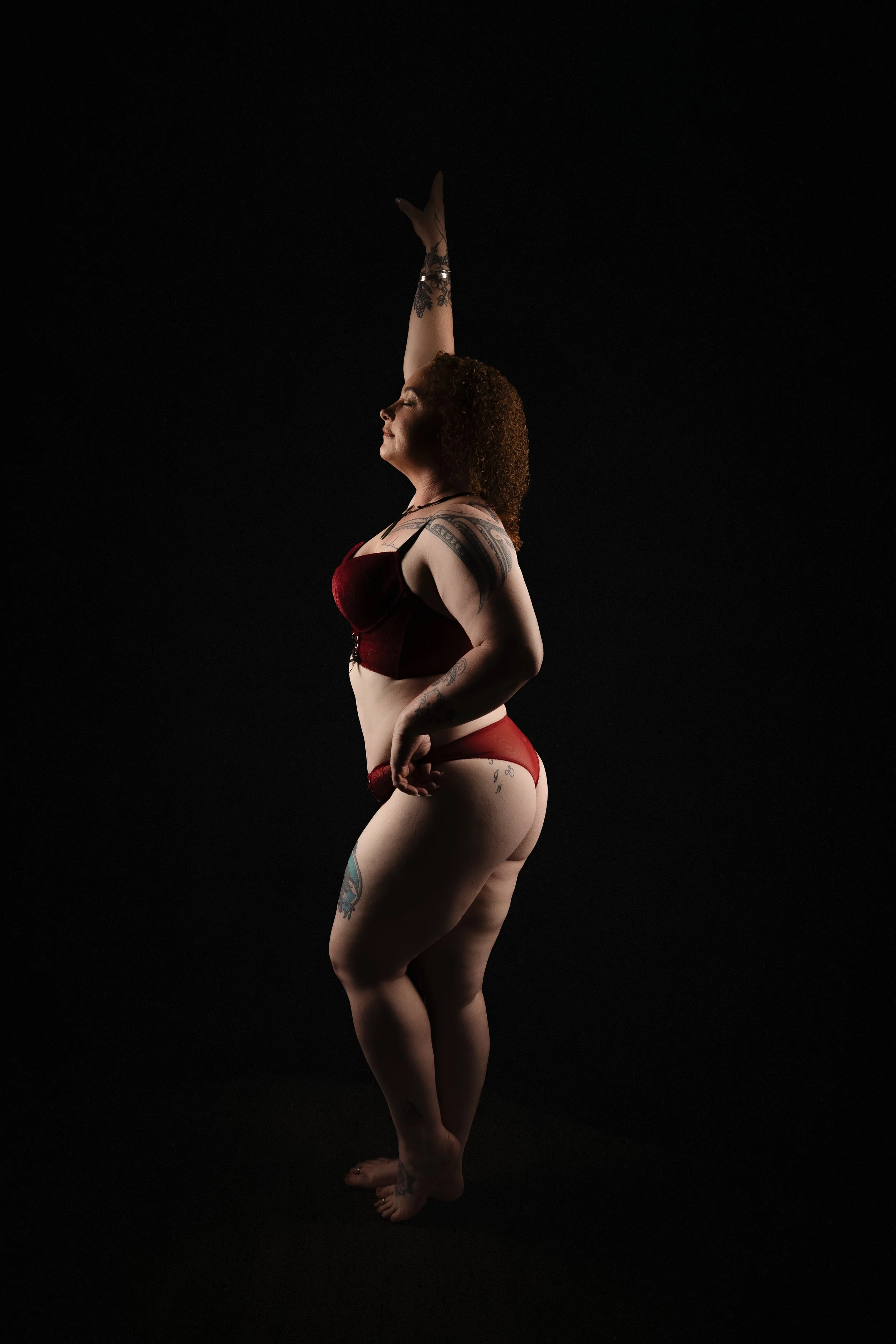 A woman with curly hair and multiple tattoos on her arm, leg, and torso, standing against a black background. She is wearing red lingerie and appears to be performing a dance pose with one arm extended upward and her eyes closed.