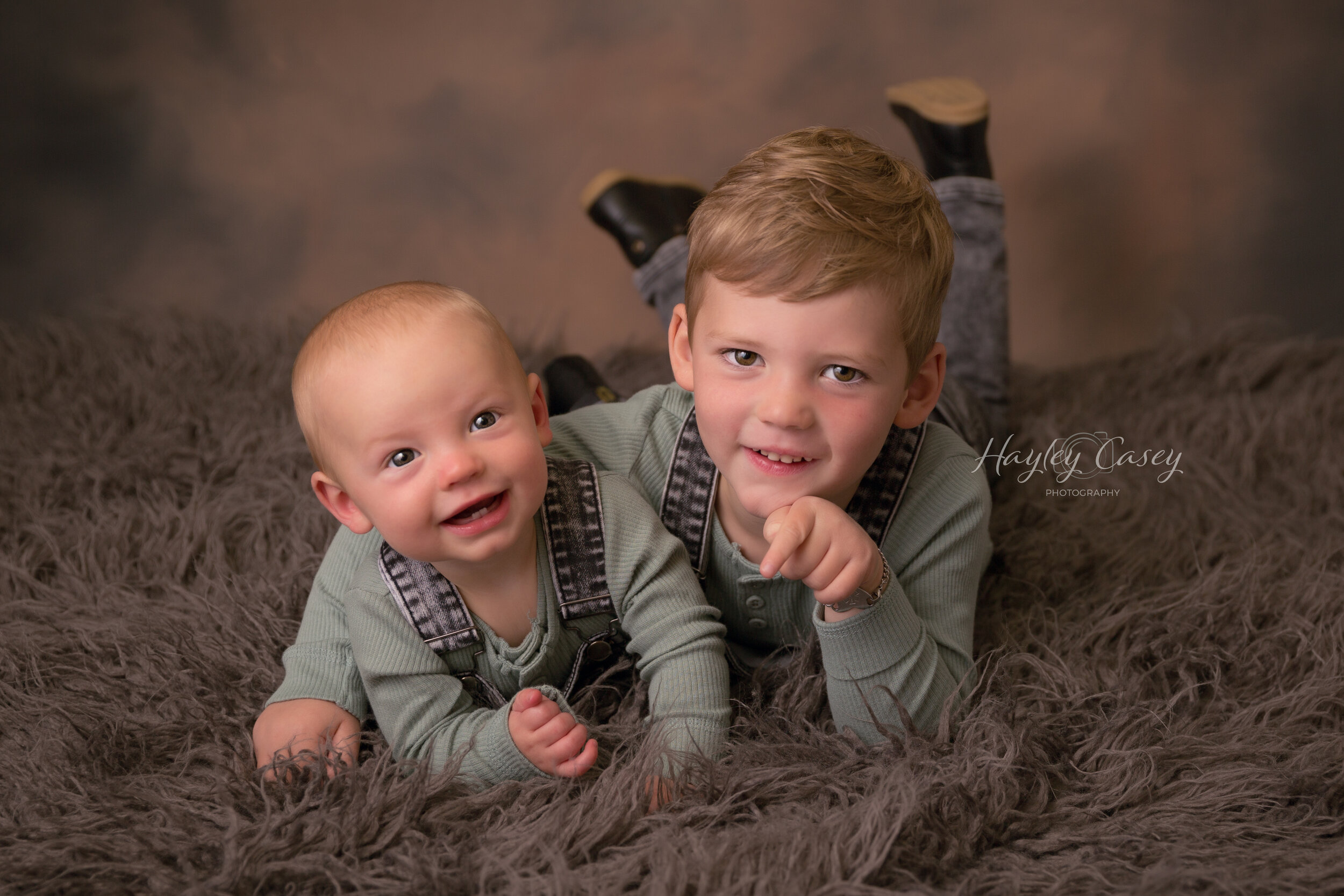 Two young children, a baby and a boy, lying on a soft gray rug indoors. They are smiling and looking at the camera, wearing matching light green outfits with dark suspenders. The boy is propped on his elbow with one hand under his chin, and the baby 