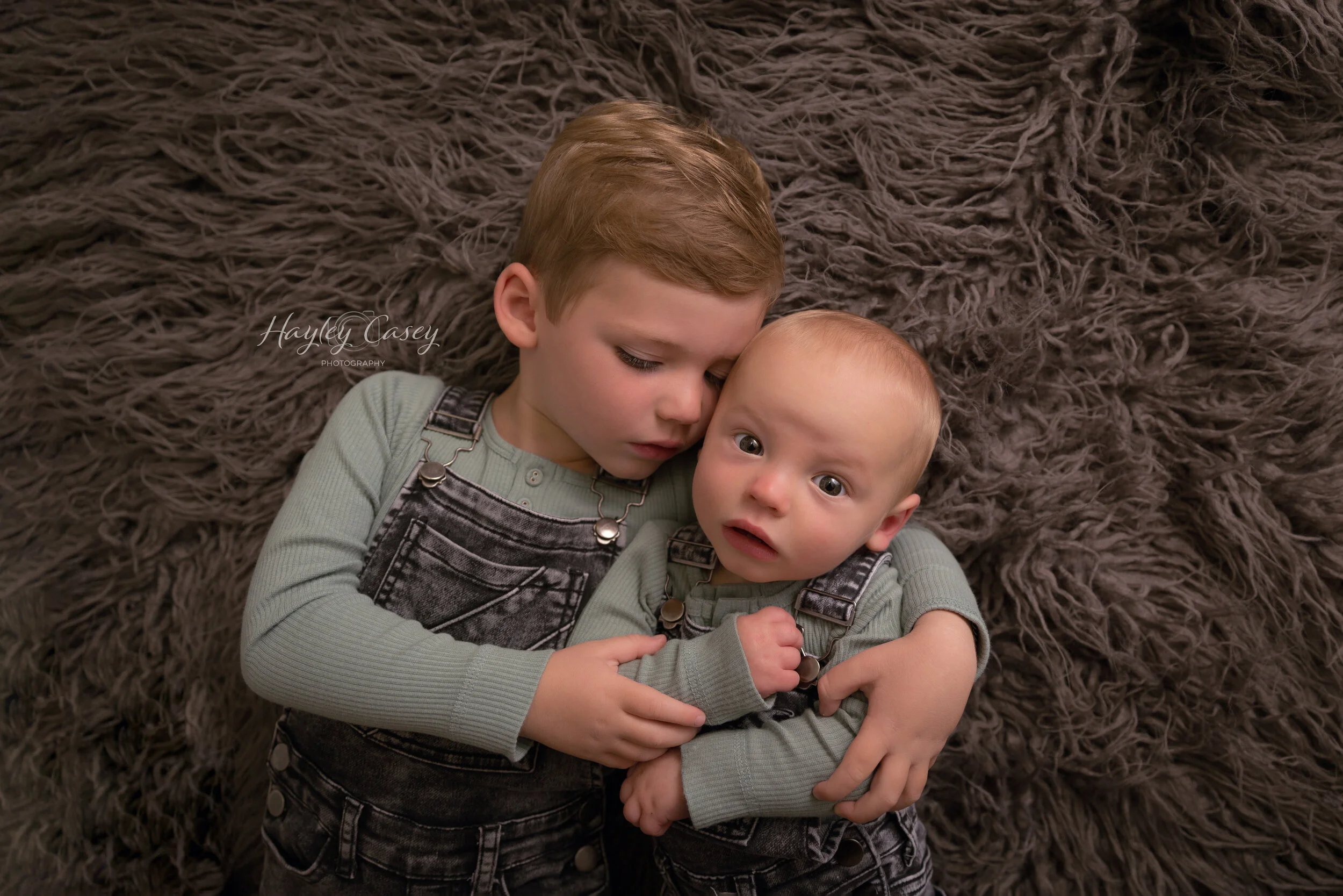 Two young boys lying on a fluffy brown rug, one holding the other close, both wearing matching green long-sleeve shirts and black denim overalls. The older boy is resting his head against the younger boy's. The younger boy has a surprised or curious 