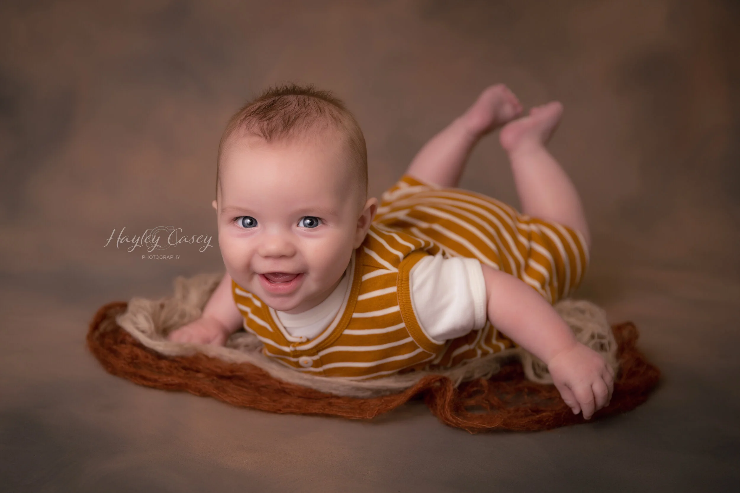 A smiling baby with bright blue eyes lying on a textured blanket on the floor, wearing a brown and white striped outfit.