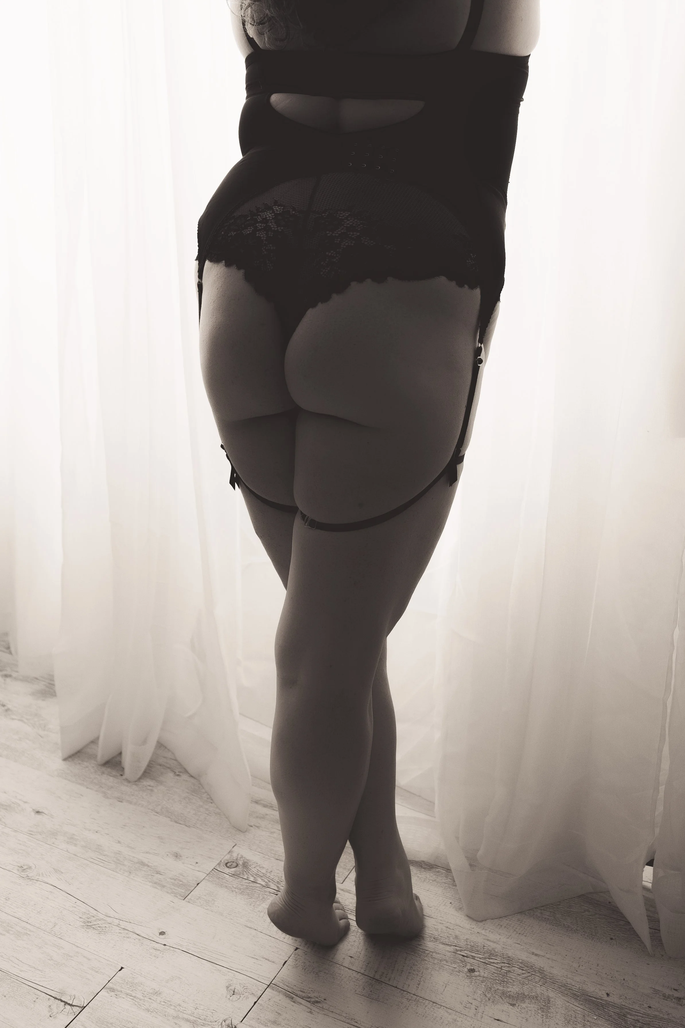 A person dressed in black lingerie with stockings and garter straps, standing with their back to the camera in front of sheer curtains letting light in.