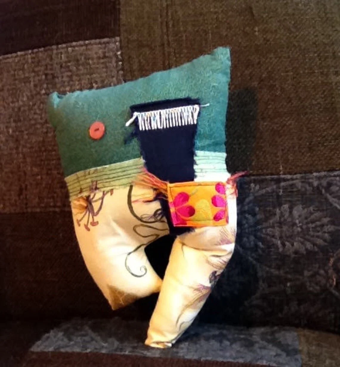 A decorative pillow with a colorful, patchwork design featuring various fabrics, buttons, and embroidered patterns, resting on a brown textured sofa.