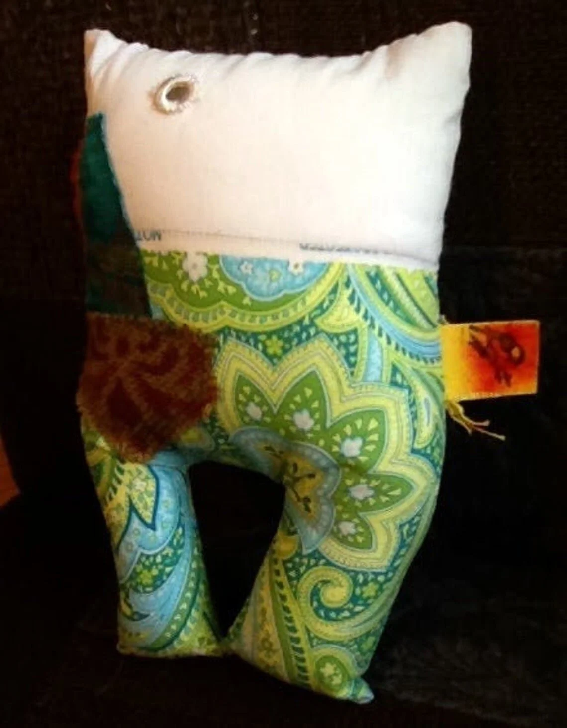A stuffed plush toy in the shape of a person with a white head, a colorful paisley-patterned body, and small fabric patches, resting on a dark surface.