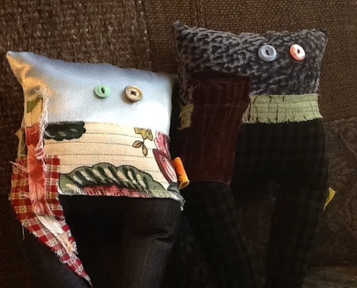Two homemade plush pillows with button eyes, made from various fabric scraps, resembling abstract creatures, placed on a brown textured surface.