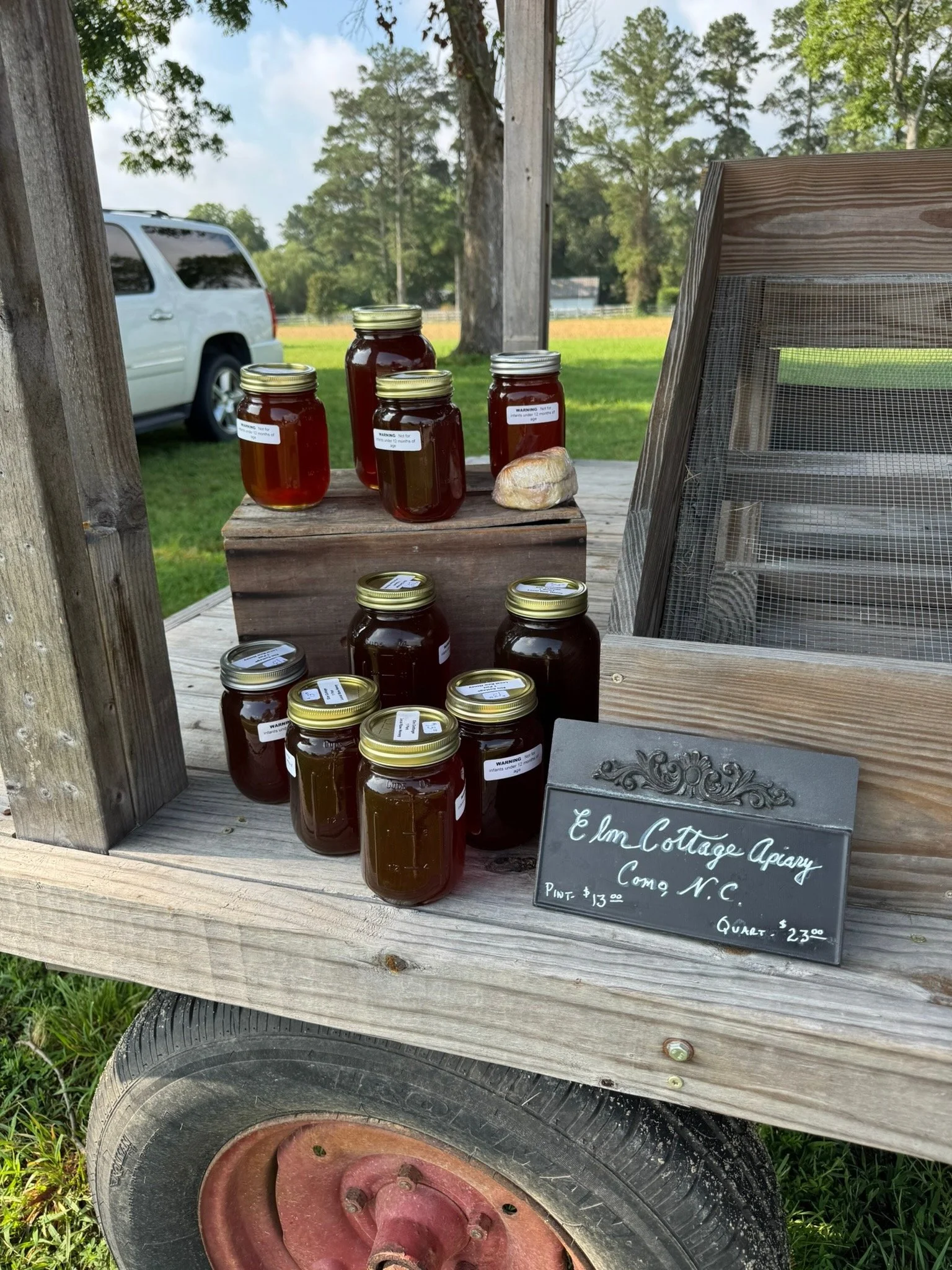 Jars of honey displayed on a wooden cart with a black chalkboard sign reading 'L&Lm Cottage Apiary, Come N.C., Pint - $13.00, Quart - $23.00.' A white vehicle is parked in the background amidst green trees and a grassy field.