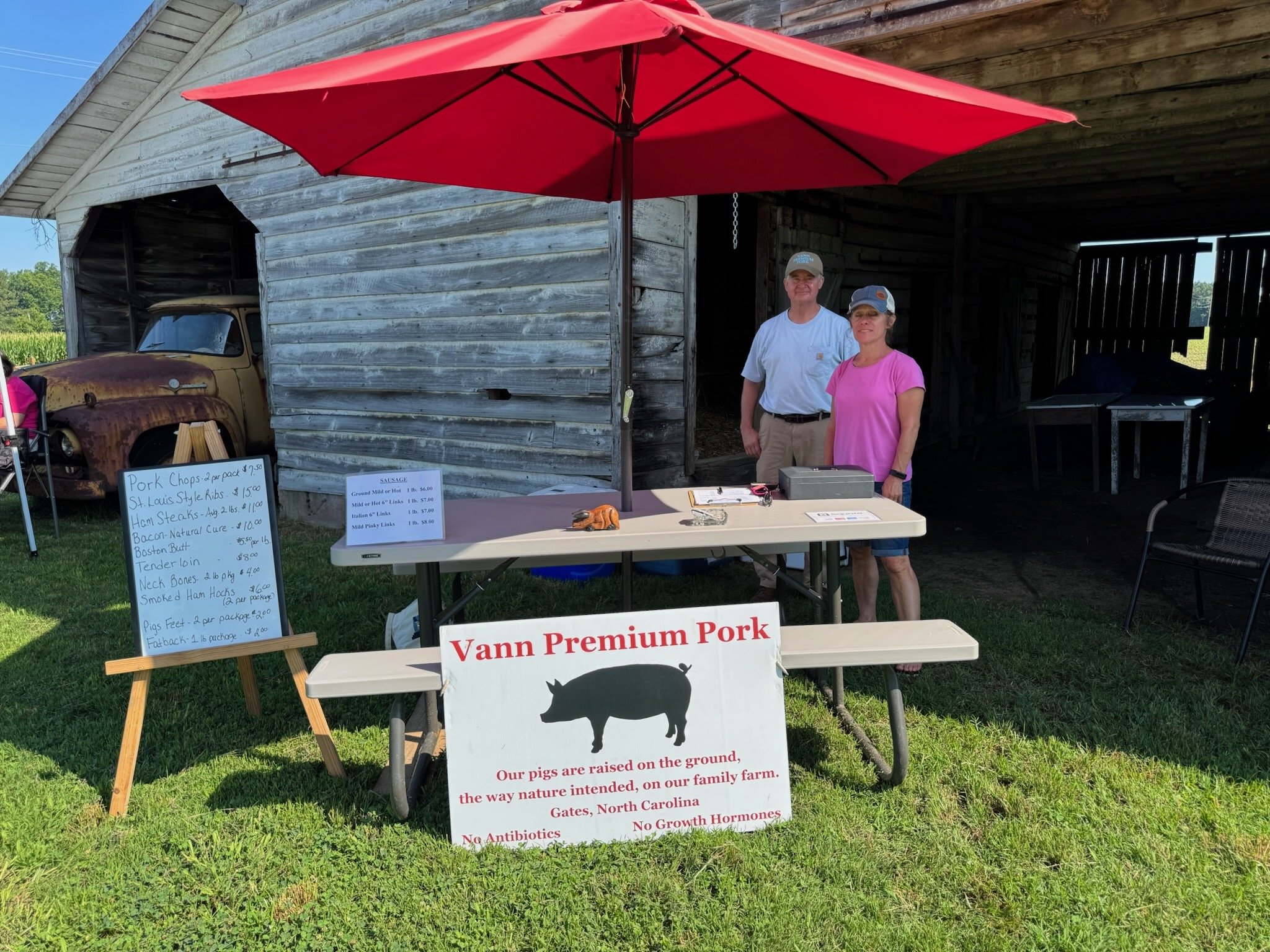 A farmers market stand with a red umbrella, two smiling people, and signs displaying various meats for sale, including pork chops, ribs, steaks, and bacon, with a rustic barn and an old rusty truck in the background.