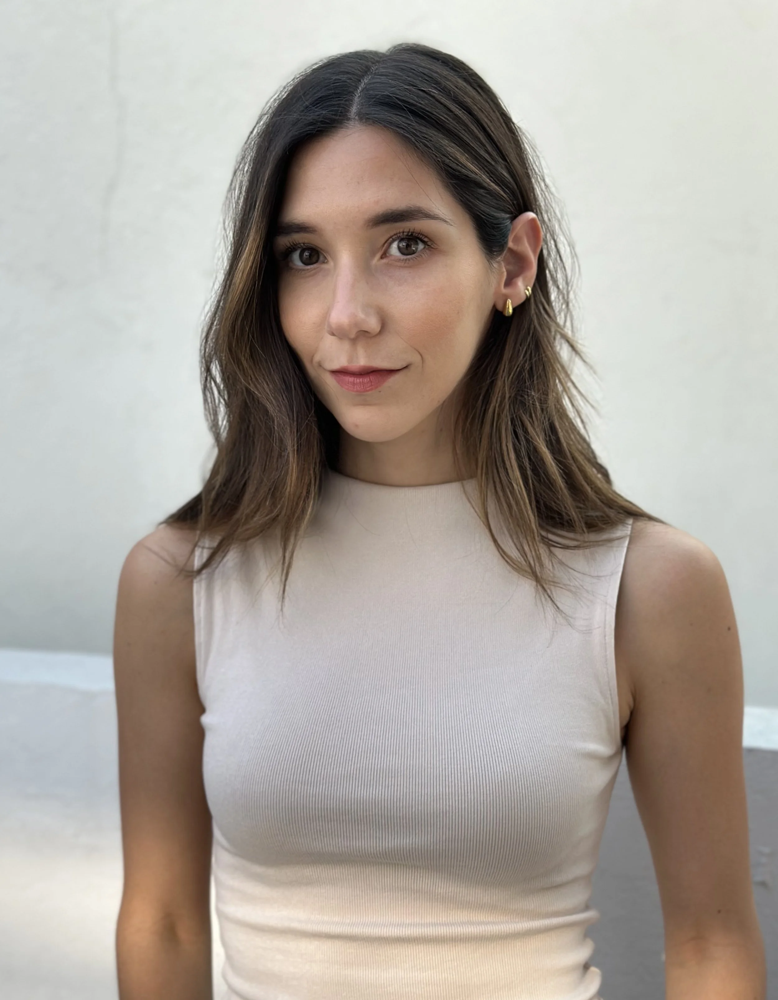 A young woman with shoulder-length brown hair and light skin, wearing a sleeveless beige top and gold earrings, standing outdoors against a plain light-colored wall.
