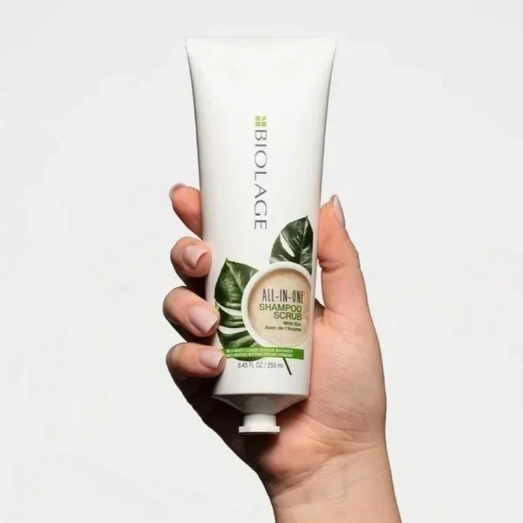 Hand holding a tube of Biolage All-in-One Shampoo and Scrub with green leaves on the label, against a plain white background.