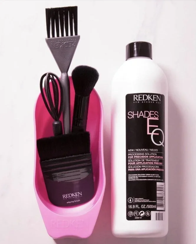 Set of hair styling tools, including a teasing comb, a hairbrush, and a silicone spatula in a pink holder, next to a bottle of Redken Shades EQ hair processing solution.