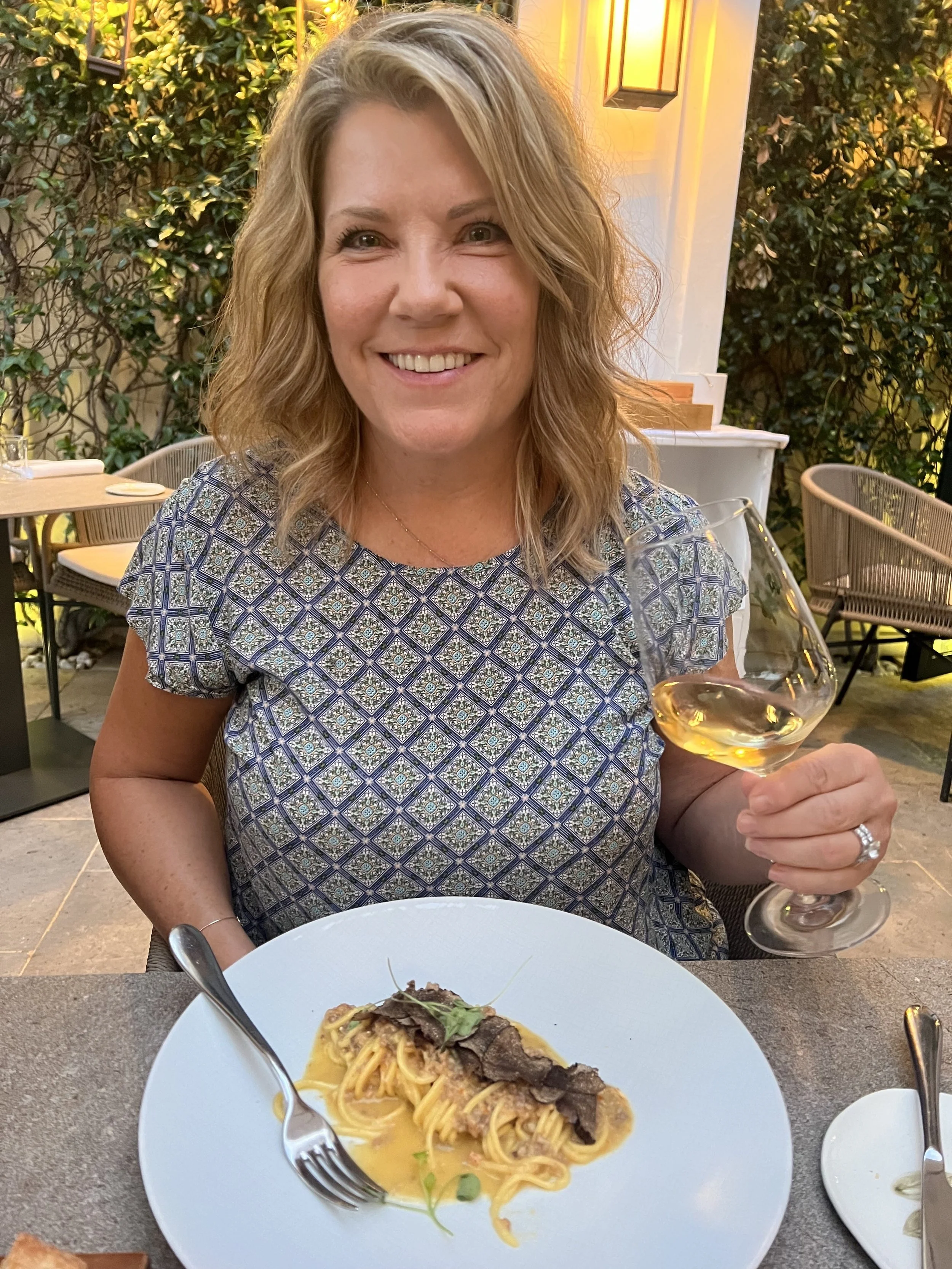 A woman smiling and holding a glass of white wine while sitting at an outdoor restaurant table with a plate of pasta in front of her.