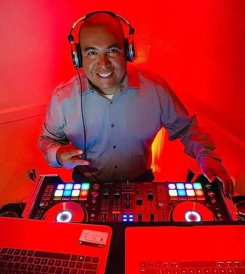 A DJ wearing a fedora hat, white shirt, and dark vest, operating a DJ controller outdoors surrounded by trees and greenery.
