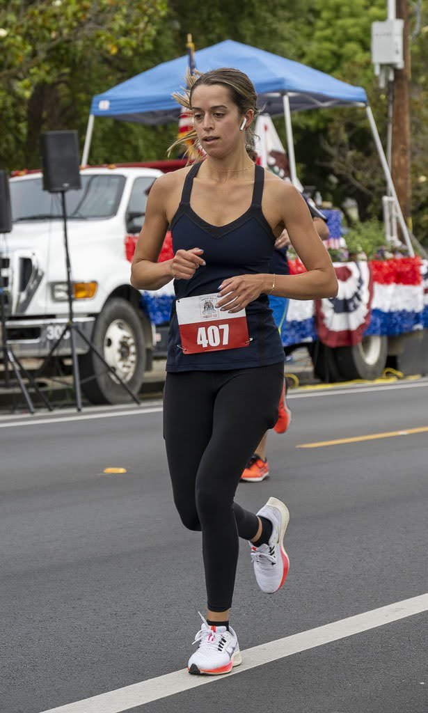 Female runner wearing black athletic attire and white running shoes participating in a race on a city street, with a parade float decorated with red, white, and blue bunting in the background.