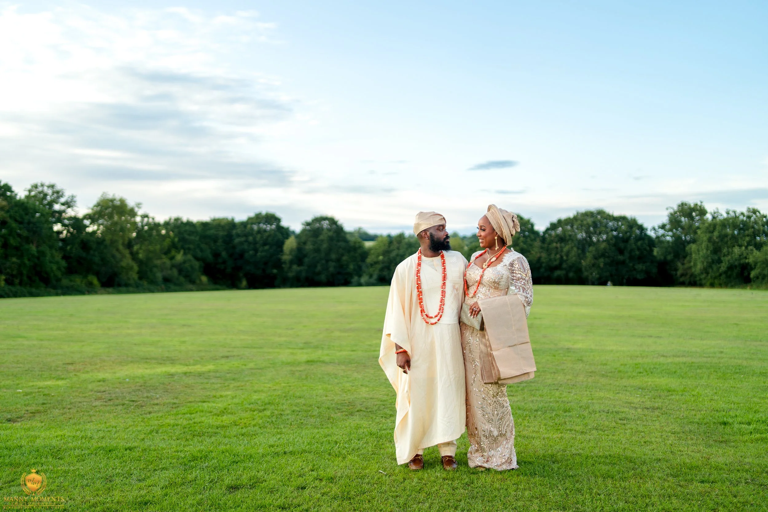 A couple dressed in traditional African wedding attire standing in a green field with trees in the background, facing each other with happy expressions.