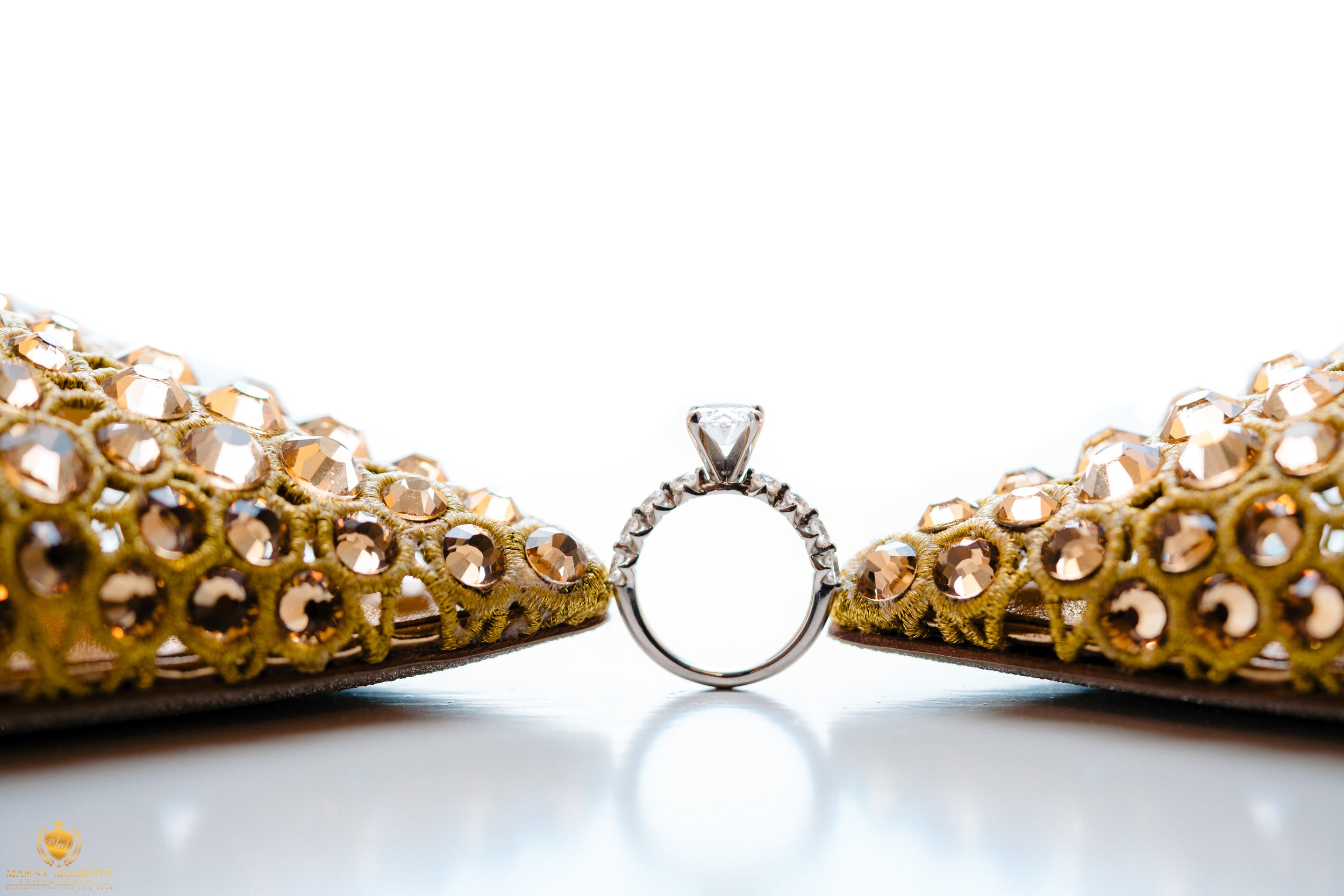 A close-up of a diamond engagement ring placed between a pair of ornate, gold-colored sandals decorated with rhinestones.