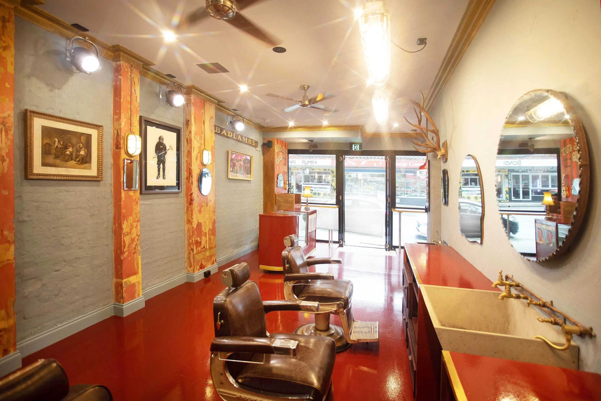 Interior of a vintage-style barber shop with red flooring, brown leather chairs, mirrors on the walls, and artwork including a framed photo and a statue of a soldier. Bright lights and a ceiling fan are seen, with large windows at the entrance letting in natural light.