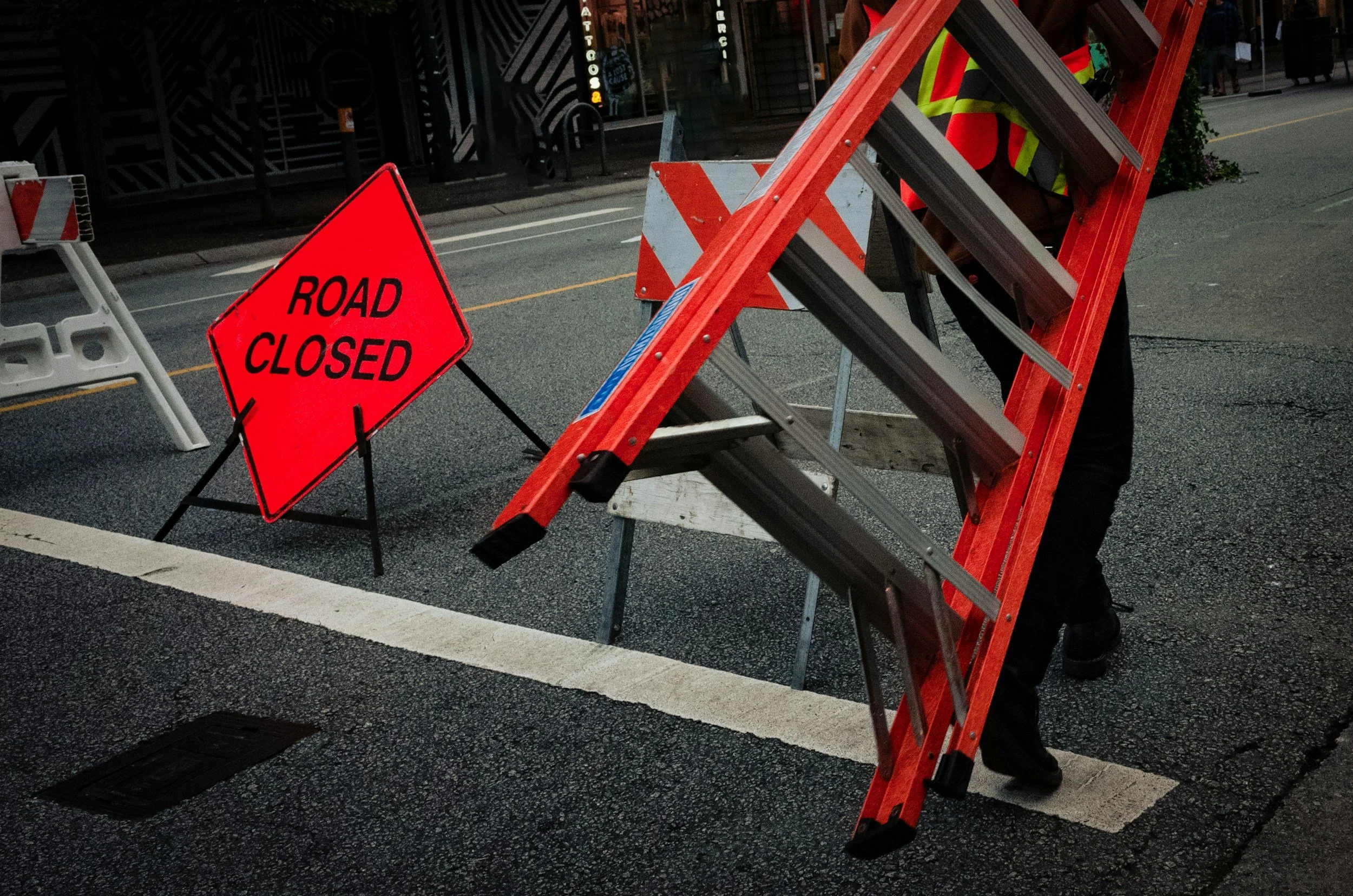 A person in a yellow safety vest working behind a wooden ladder on a closed street, with a red 'Road Closed' sign in the foreground.