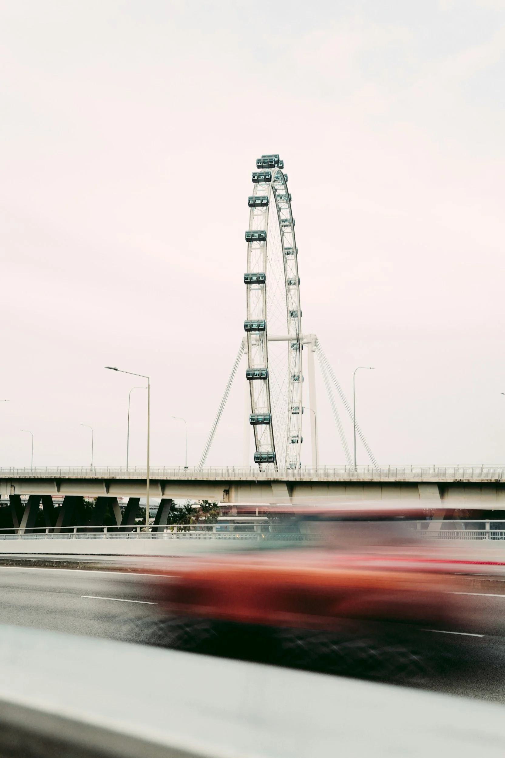 A large Ferris wheel viewed from a moving vehicle on a highway with blurred red car passing by. The Ferris wheel is against a cloudy sky.