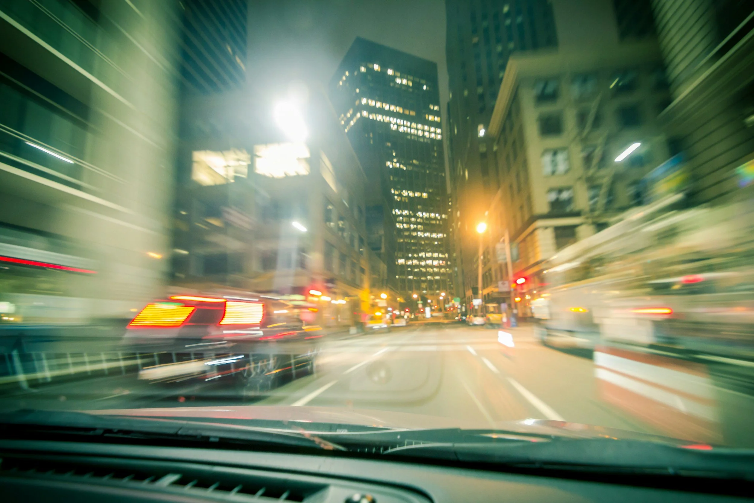 Nighttime city street view from inside a moving vehicle with blurred lights and buildings.