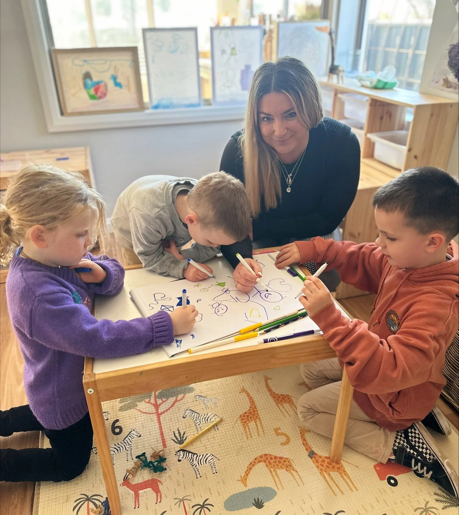 Children and an adult drawing on a large sheet of paper at a small table in a classroom setting.