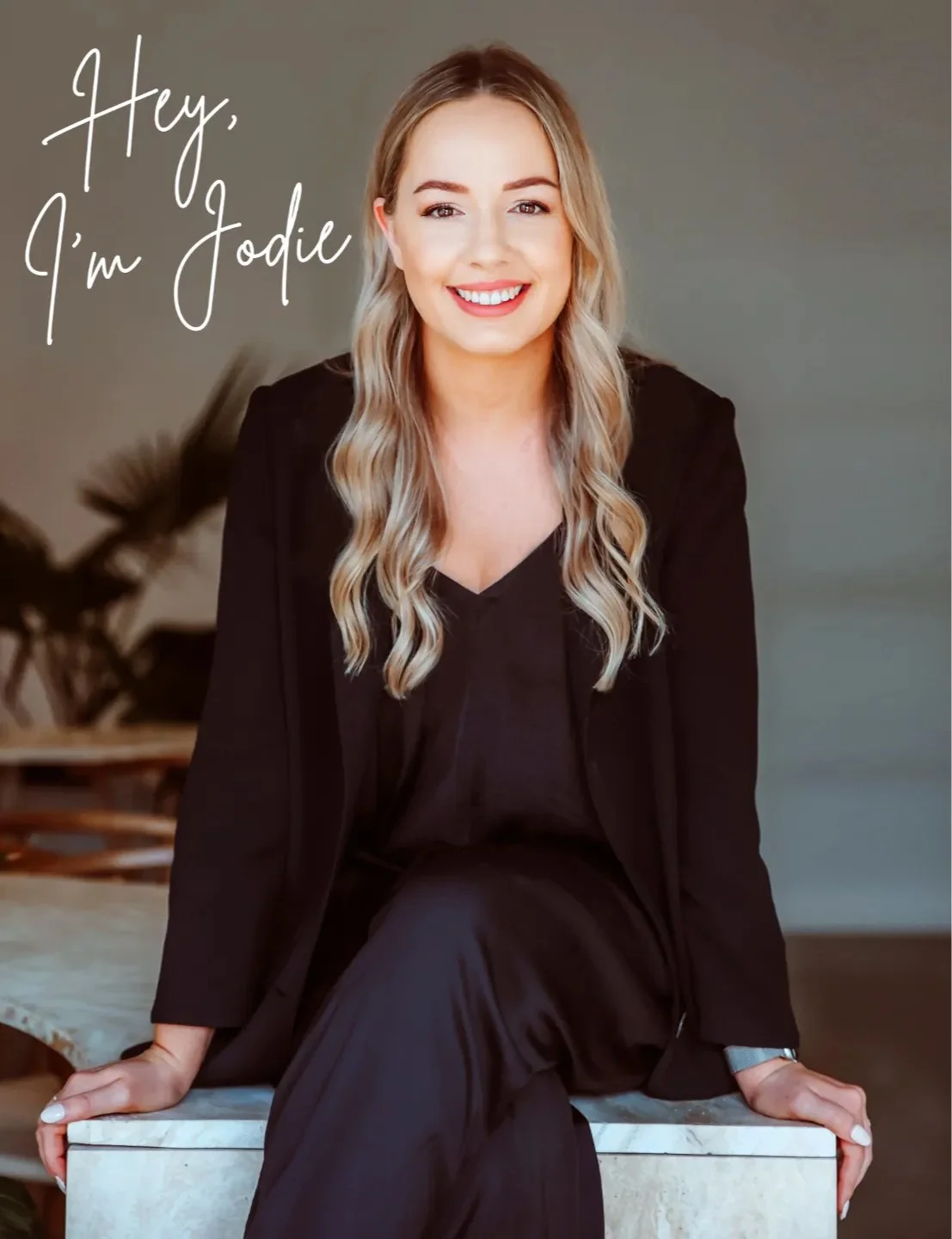 Smiling woman with blonde hair in a black outfit, sitting on a marble surface, with "Hey, I'm Jodie" text.