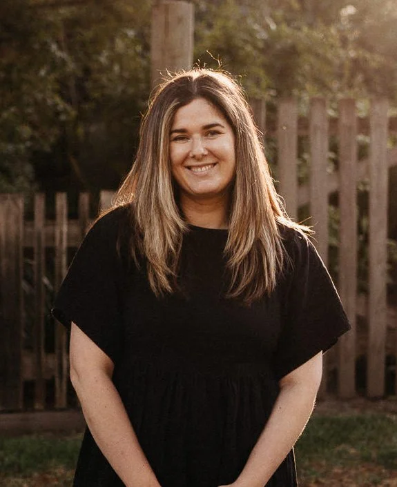 Smiling person in black dress standing outdoors near a wooden fence with greenery in the background.