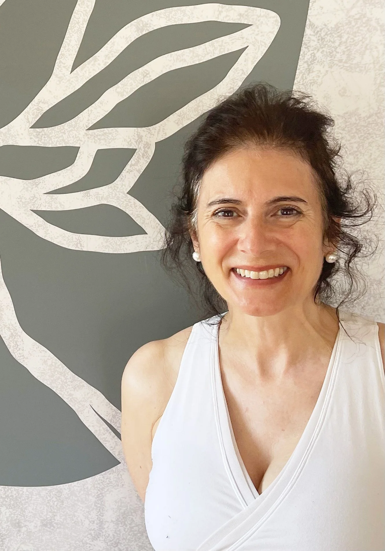 Smiling woman in a white top standing in front of a gray wall with abstract leaf design.