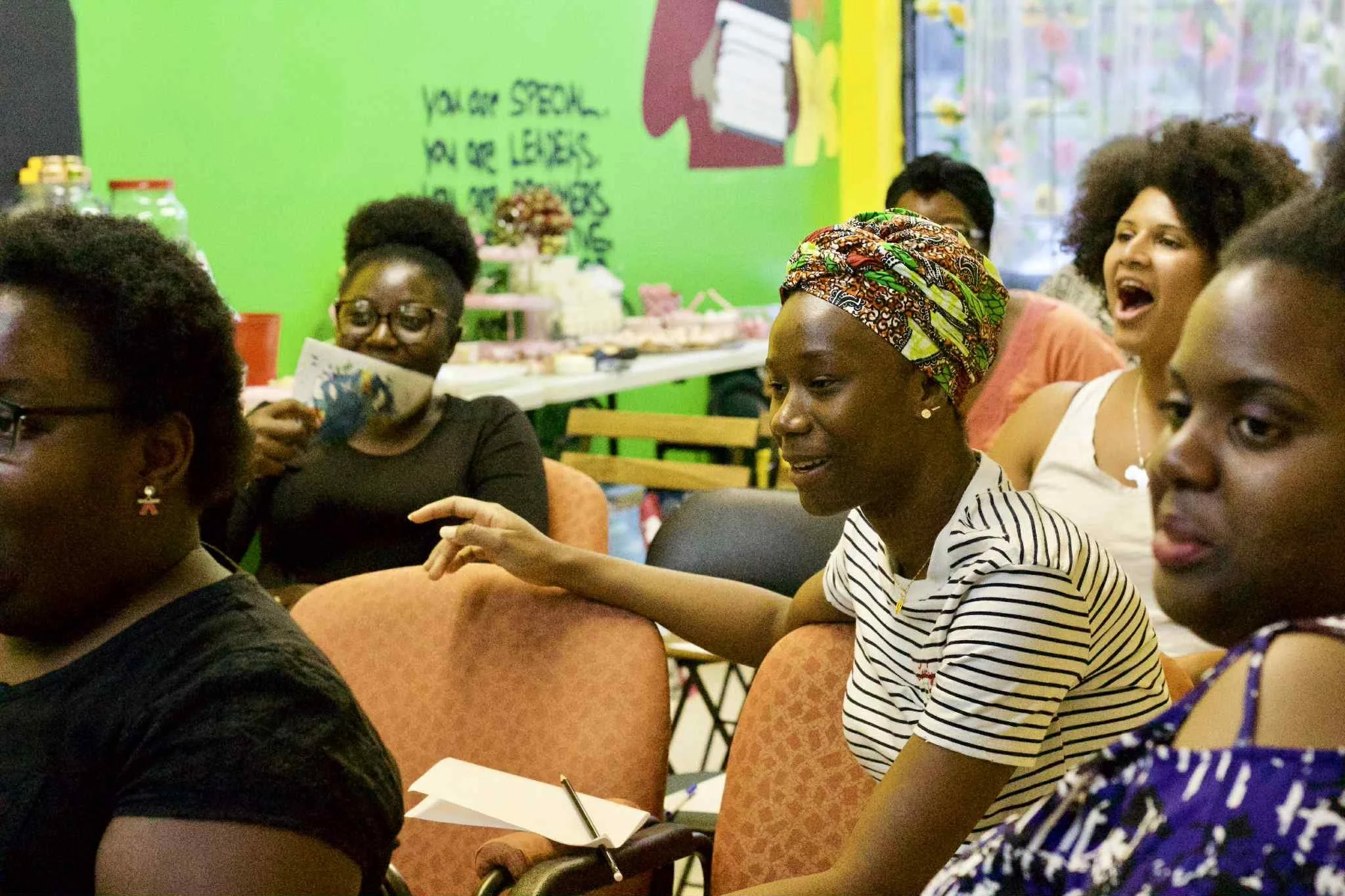 Group of women sitting and engaging in conversation at a colorful indoor event with a bright green wall in the background.