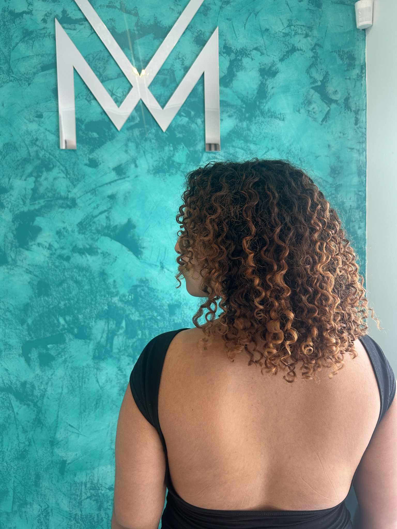 A woman with curly hair standing with her back to the camera, facing a teal textured wall with a metallic WWE logo on it.