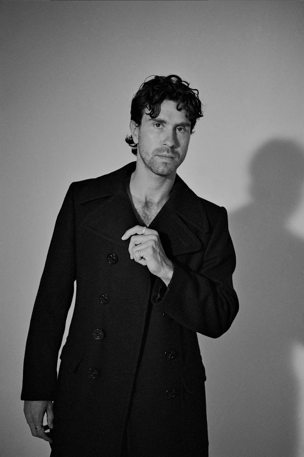 Black and white portrait of a man with wavy hair, wearing a dark, double-breasted coat, standing against a plain background.