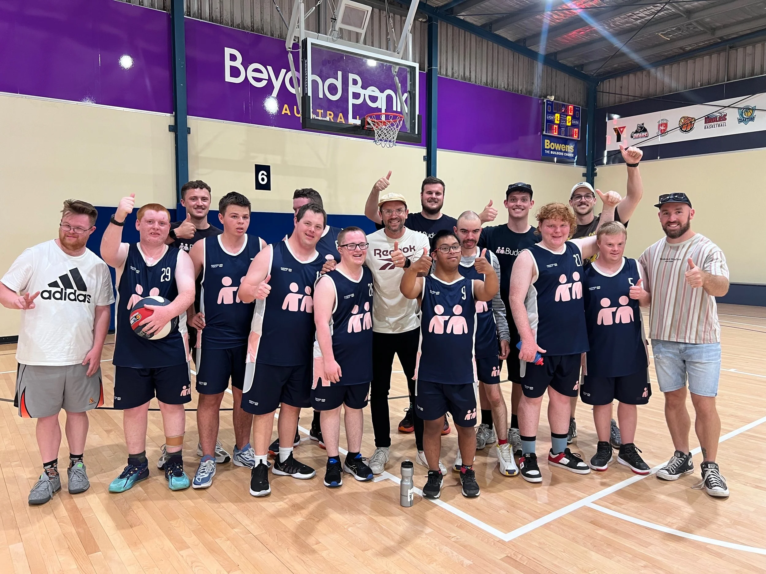 A group of volleyball players and coaches posing for a photo in an indoor volleyball court, some holding a ball and giving thumbs up.