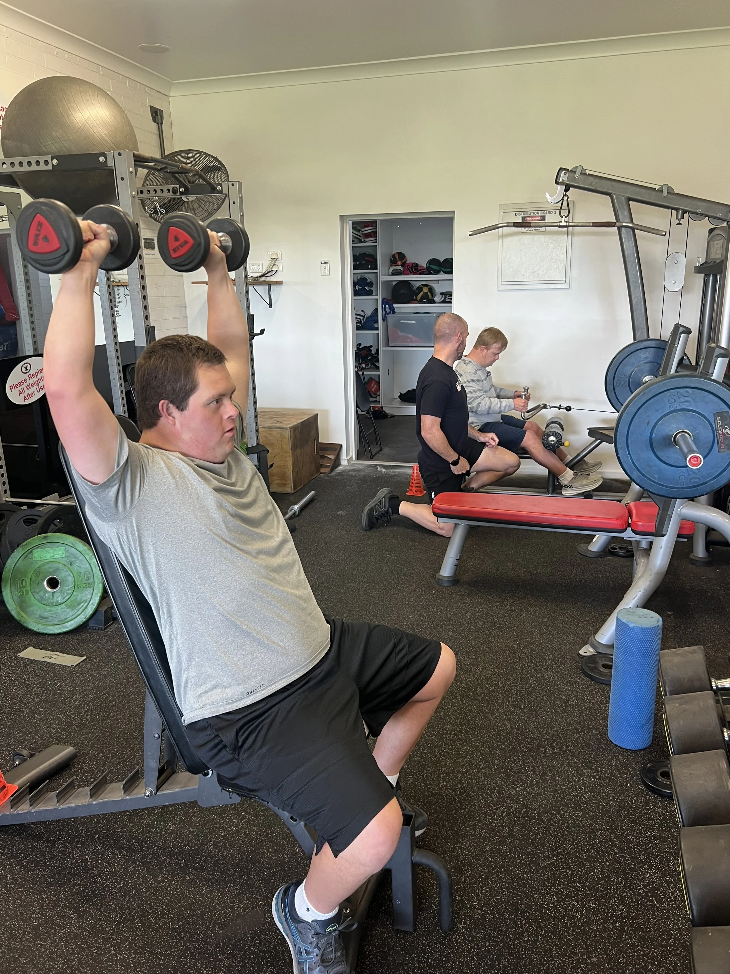 Three men exercising in a gym. The man in the foreground is lifting dumbbells while seated. The two men in the background are using a rowing machine.