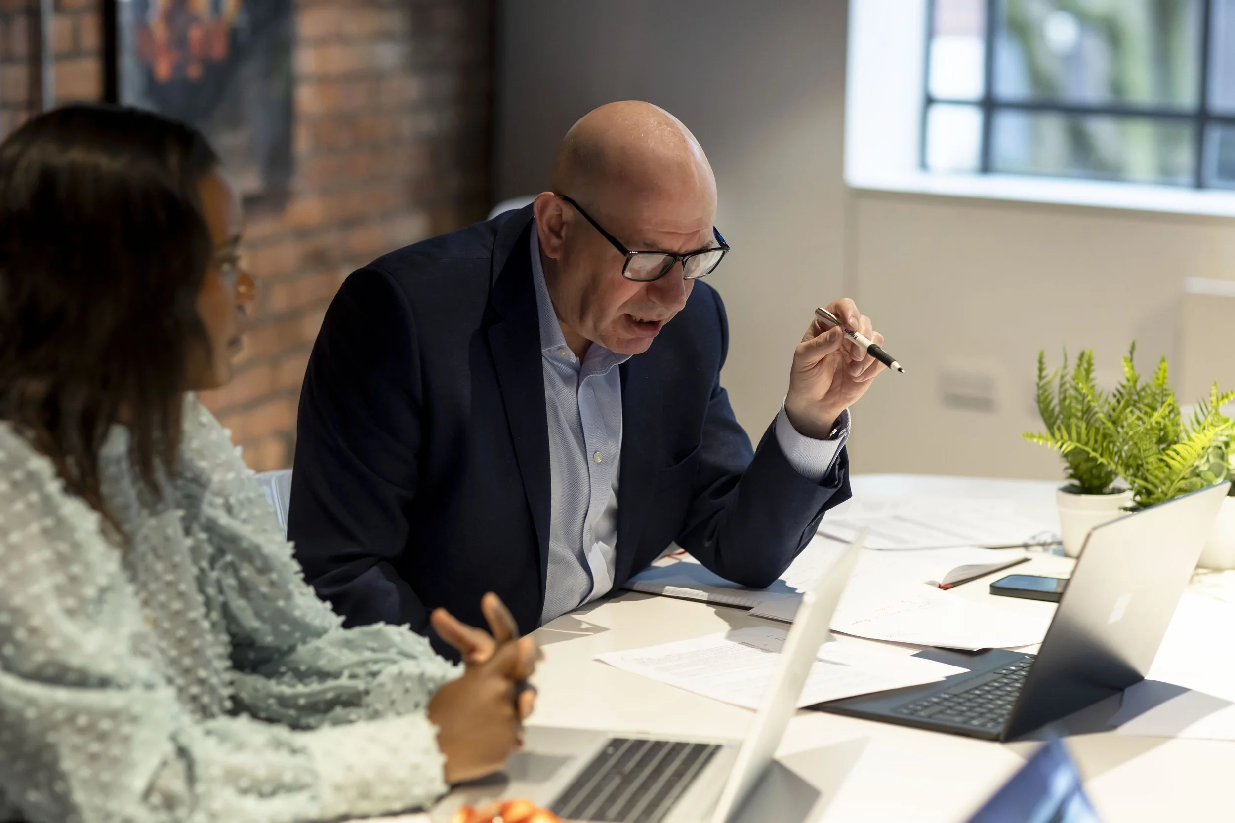 Two people engaged in a discussion at a conference table in an office setting, with laptops, papers, and a potted plant visible.