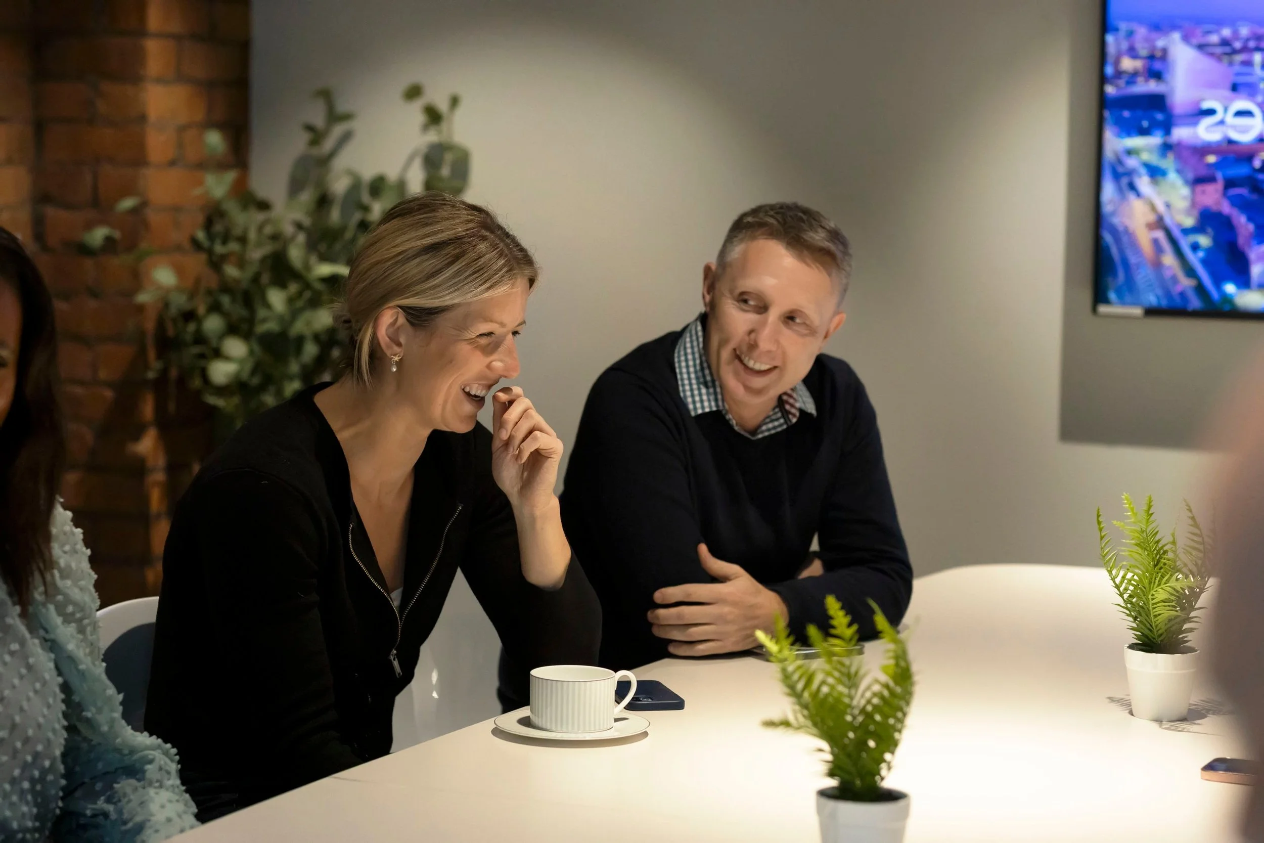 Two people sitting at a table, smiling and laughing during a meeting or casual conversation; a woman with short blonde hair wearing a black top, a man with short light brown hair in a dark sweater with a checkered collared shirt underneath; a coffee cup on a saucer, potted plants, and a cityscape | office or conference room setting.