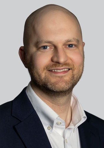 A man with a bald head and beard, wearing a white shirt and black suit jacket, smiling at the camera against a plain light background.