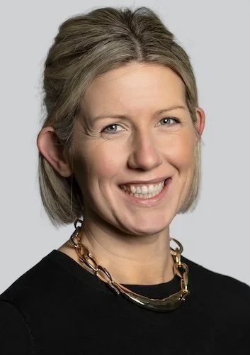 A professional woman with blonde hair wearing a dark blazer and patterned blouse, smiling lightly at the camera.