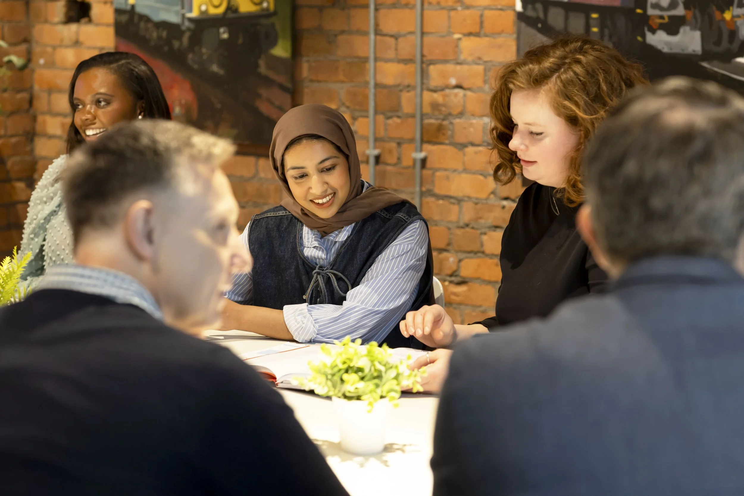 A diverse group of five people, three women and two men, sitting around a table with open books, in a cozy restaurant with brick walls, engaged in a discussion or activity, with a small plant on the table.