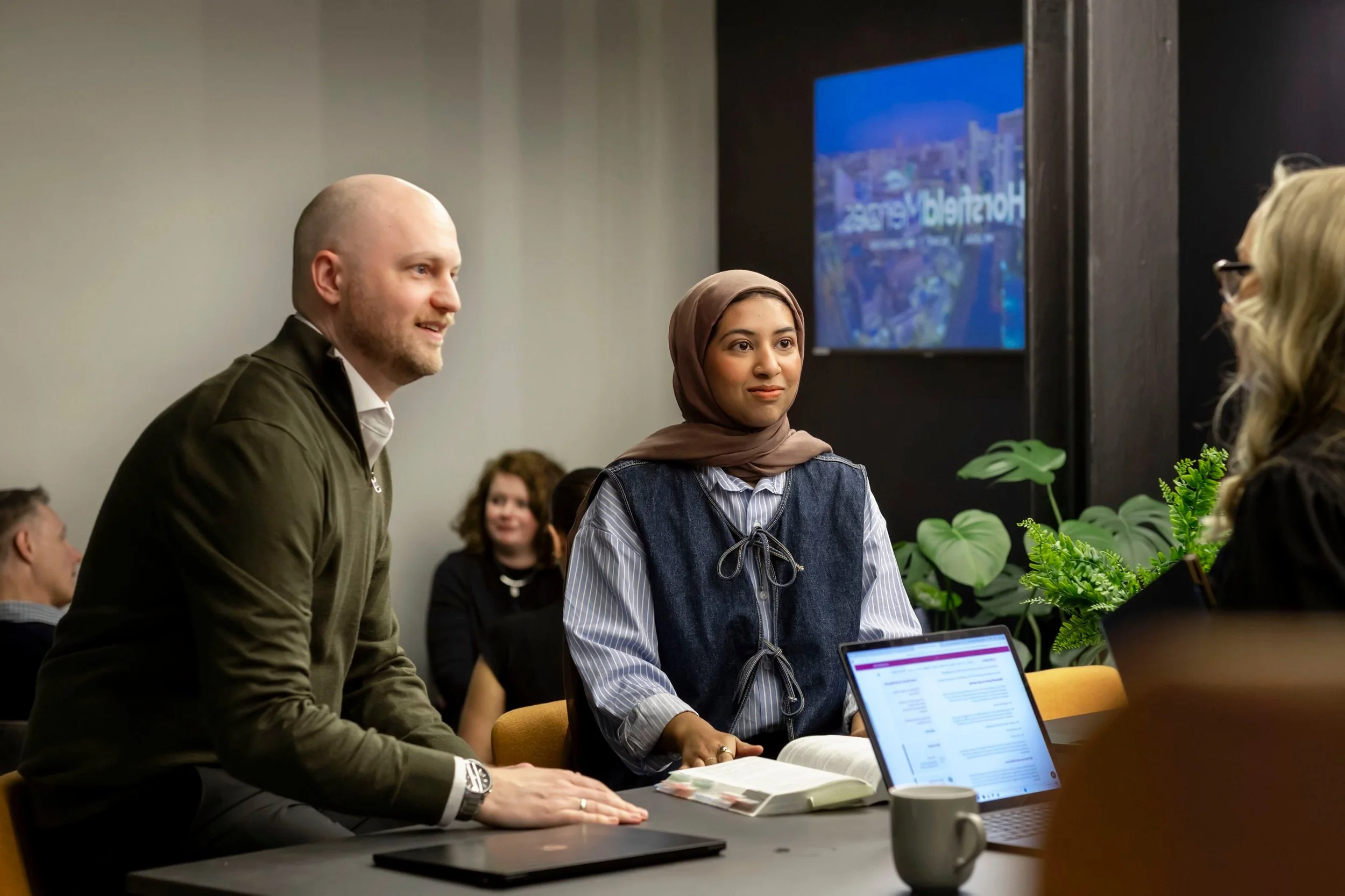 A diverse group of people engaged in a meeting or discussion in a conference room, with plants and a monitor displaying a cityscape background.