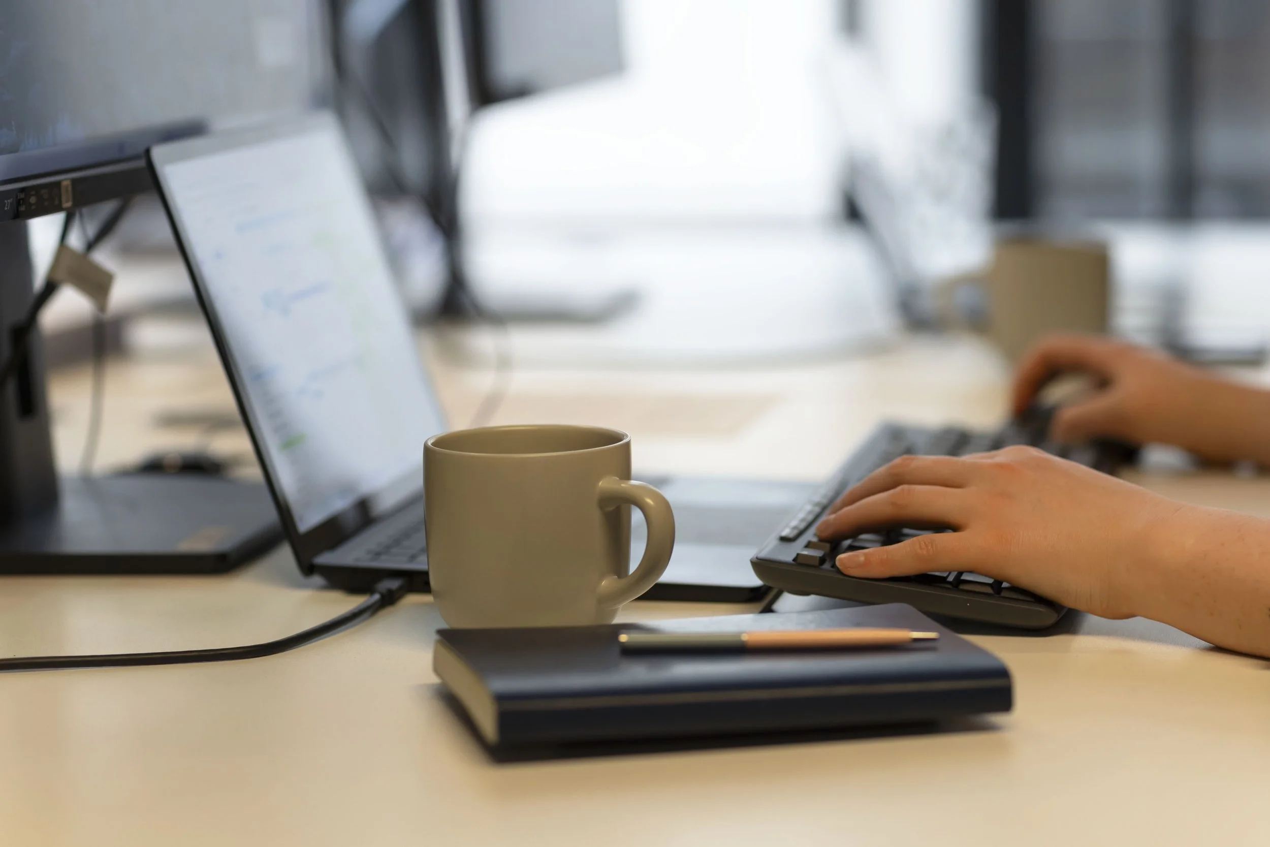 Person using a computer keyboard at a desk with a coffee mug, a closed notebook, and a pen, in an office setting.