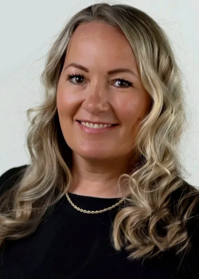 A woman with long, wavy blonde hair, wearing a black top and a gold necklace, smiling at the camera against a plain white background.