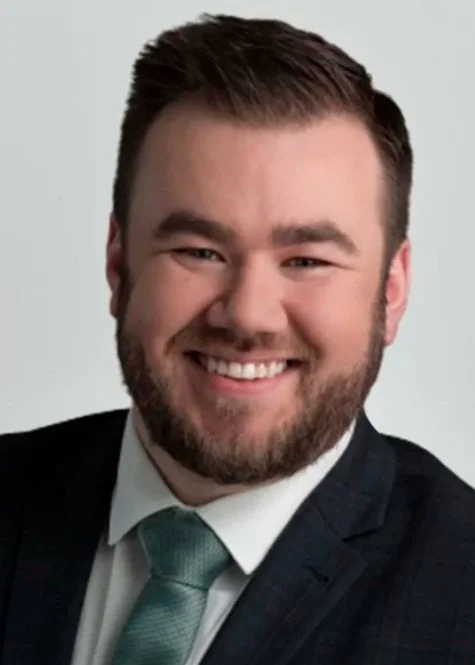 A smiling man with brown hair and a beard wearing a dark suit, white shirt, green tie, and pocket square, posed against a light gray background.