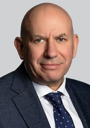 A bald man in a navy blazer and light blue dress shirt, standing with arms crossed against a plain light-colored background.