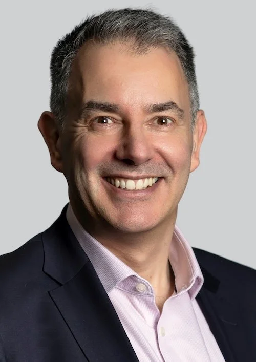 A smiling man with dark hair, wearing glasses, a white shirt, and a dark blazer, standing against a light background.