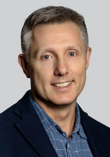 A man smiling, wearing a black suit jacket and white shirt, with short brown hair, against a plain light background.