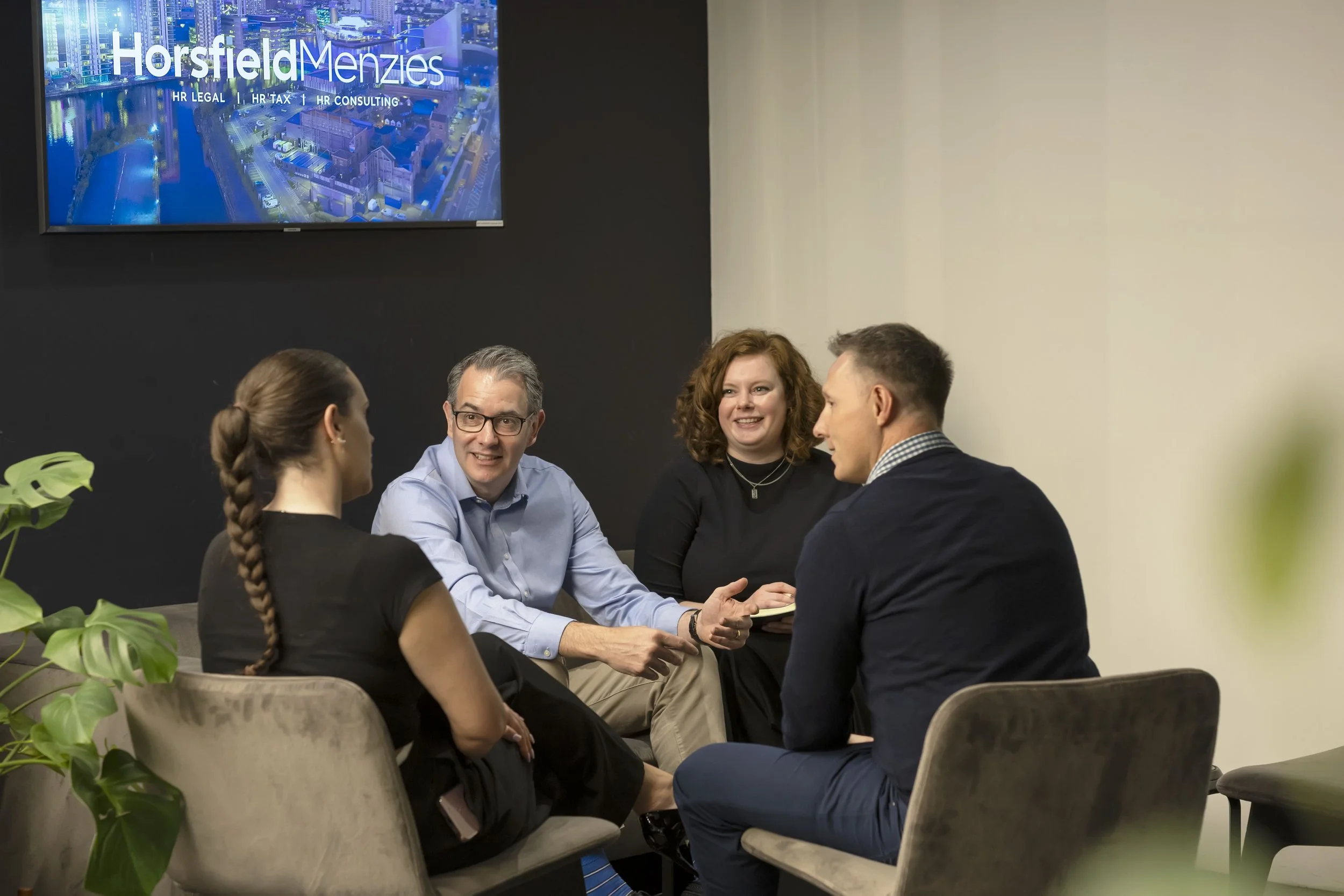 Four business professionals having a discussion in a modern office conference room, with a monitor displaying 'Horsfield Menzies' and services such as HR Legal, HR Tax, and HR Consulting in the background.