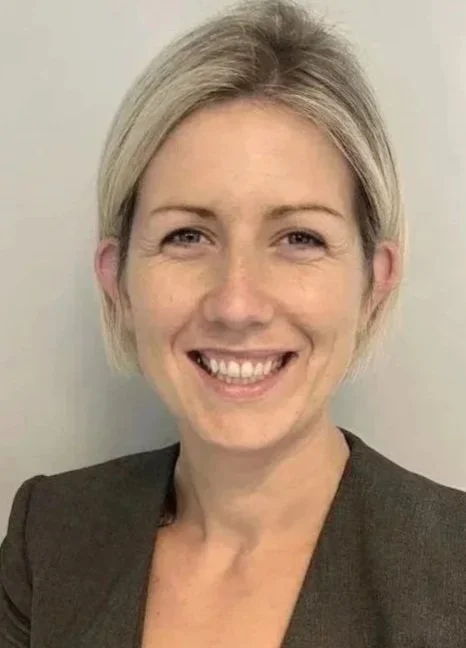 A professional woman with blonde hair wearing a dark blazer and patterned blouse, smiling lightly at the camera.