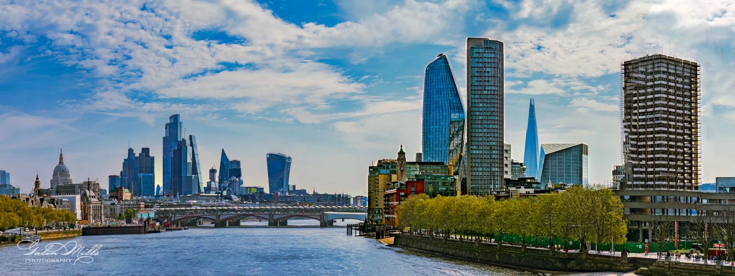 Panoramic view of London skyline with modern skyscrapers, St. Paul's Cathedral, and the River Thames under a bright cloudy sky.