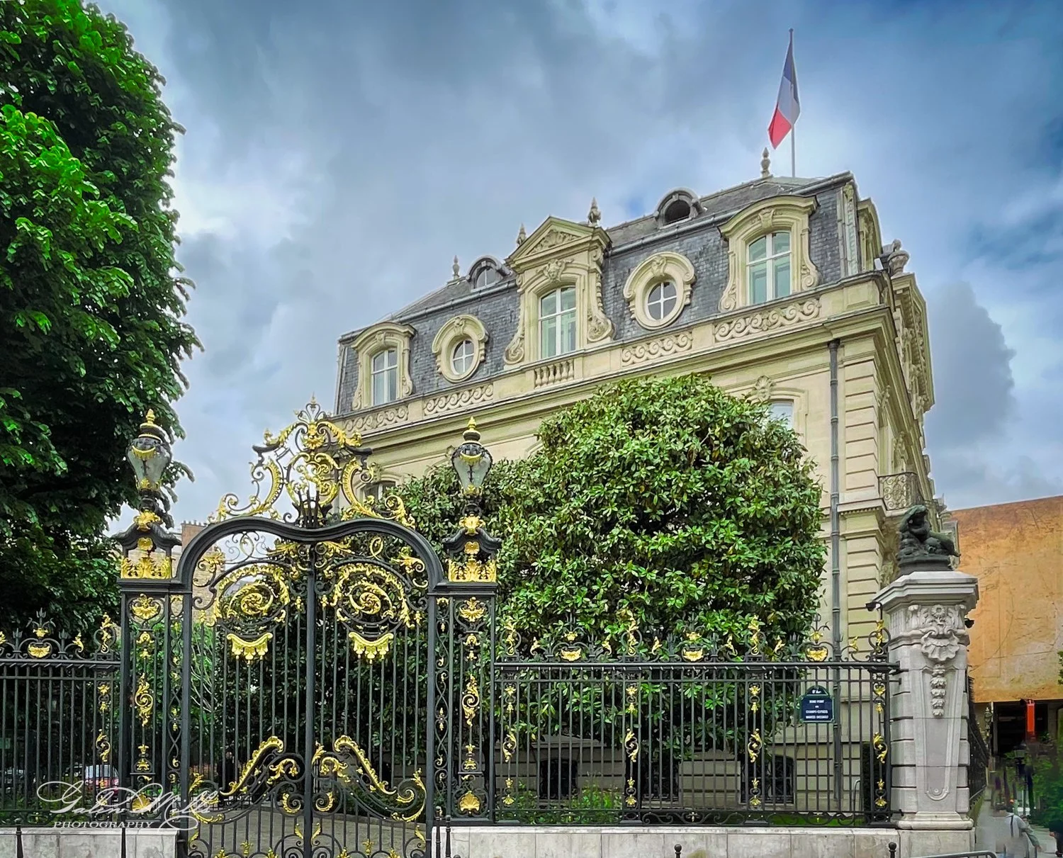 A historic building with ornate architecture and a French flag on the roof, behind decorative wrought iron gates and surrounded by lush greenery.