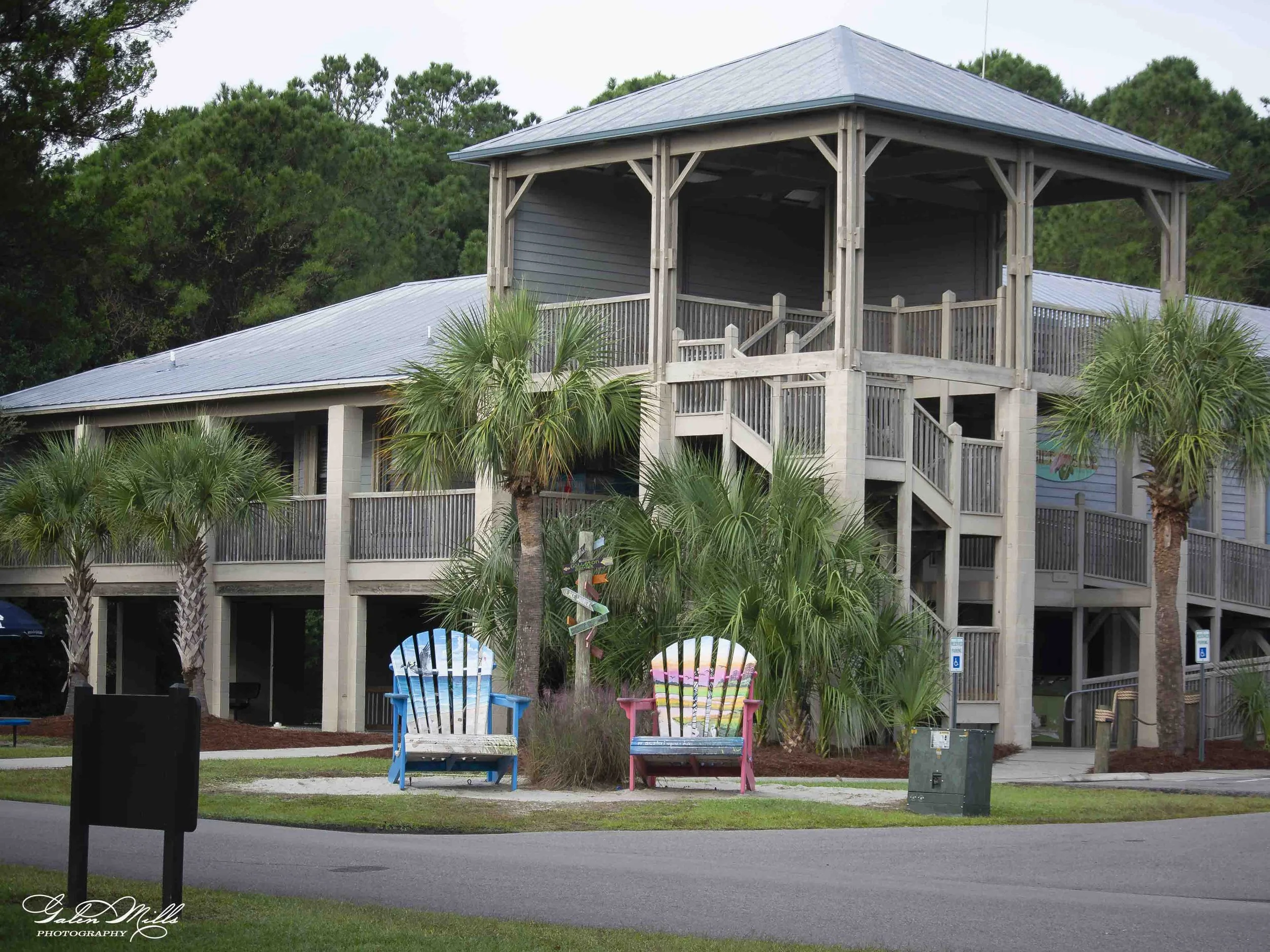 HBSP two-story wooden building with a gray metal roof, surrounded by palm trees and colorful Adirondack chairs in the foreground.