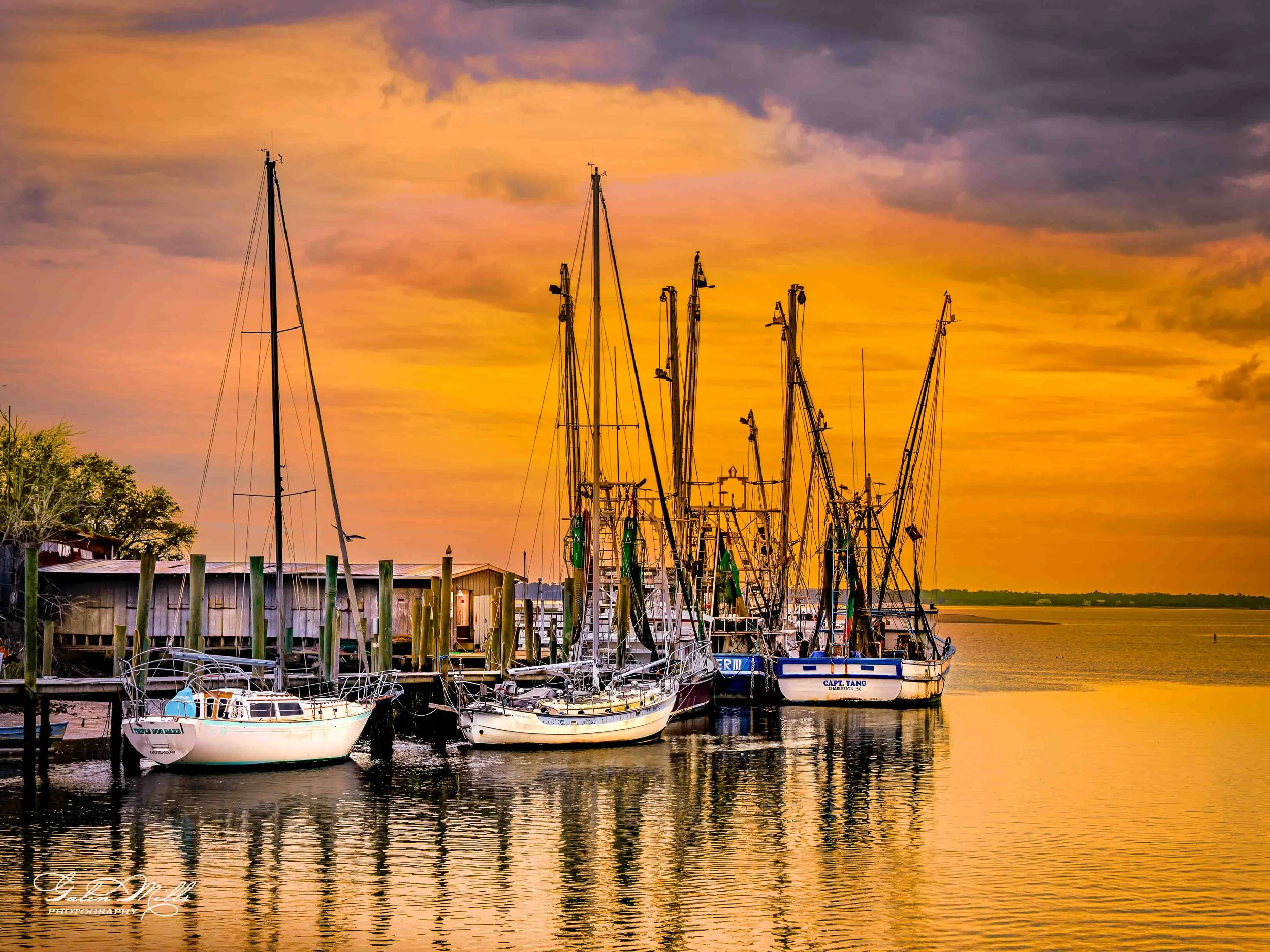 Several sailboats docked at a pier during a colorful sunset, with water reflecting the vibrant sky.